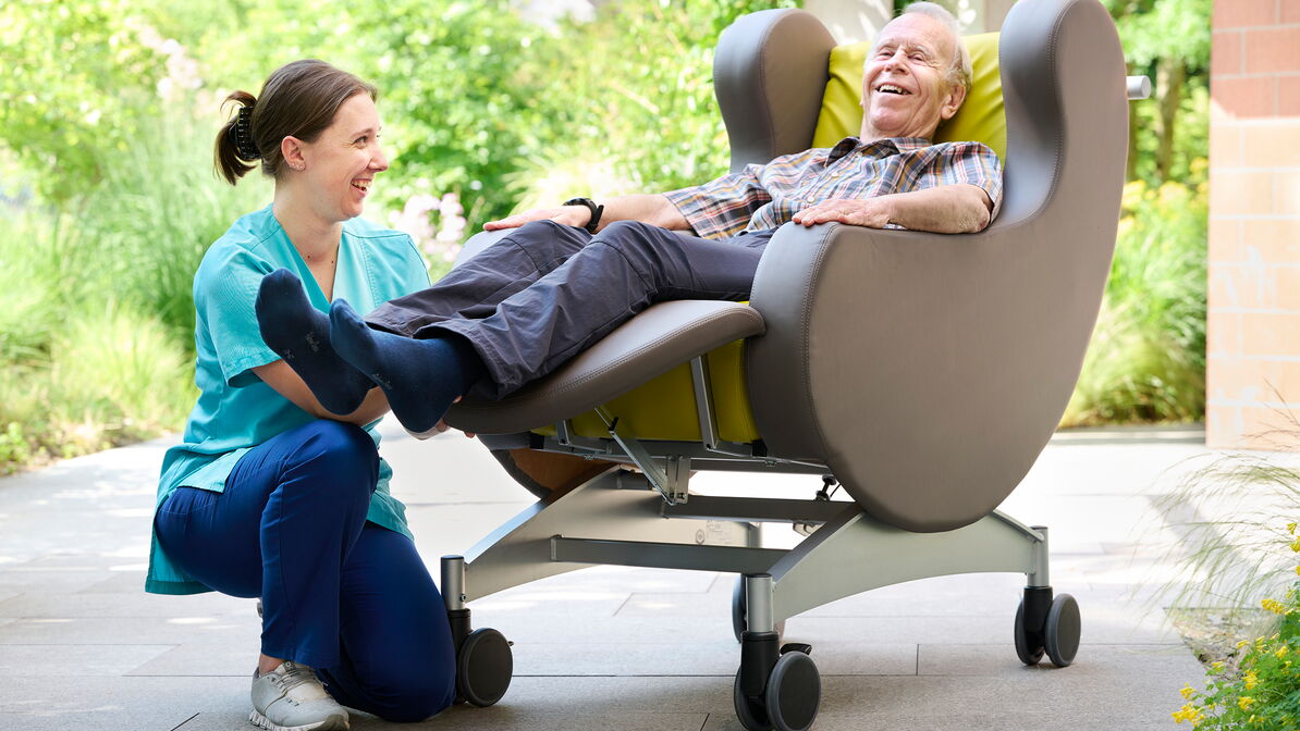 Un homme âgé est allongé de façon détendue dans un fauteuil de repos spécial, tandis qu’une professionnelle des soins est agenouillée à côté de lui et soutient ses jambes en souriant.