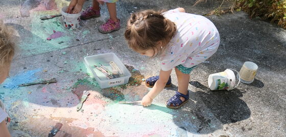 Une petite fille peint au sol avec des pinceaux et de la peinture à l’extérieur, tandis que d’autres enfants et du matériel de peinture sont visibles.