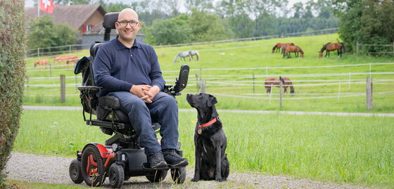 Matyas Sagi-Kiss sitzt im Rollstuhl in der Natur; neben ihm sitzt sein Assistenzhund.