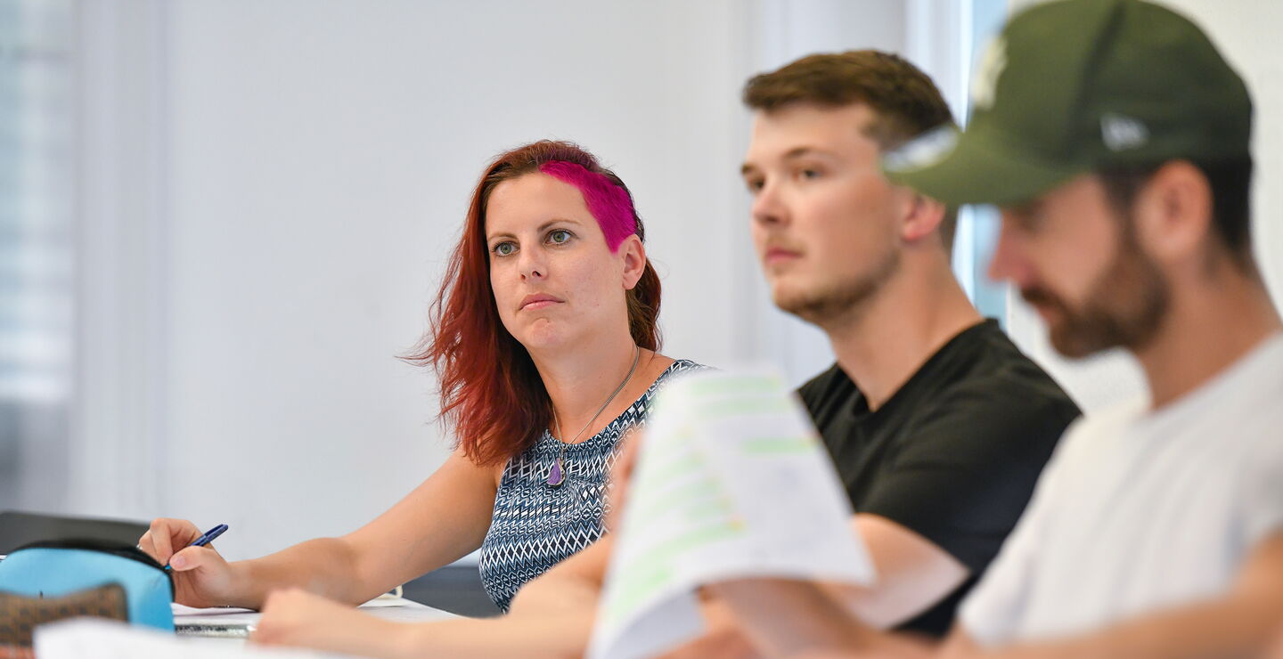 Mehrere Teilnehmende sitzen an einem Tisch in einem Unterrichtsraum, hören zu und arbeiten mit Unterlagen.