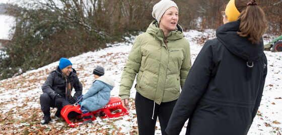 Zwei Erwachsene sprechen im Freien im Schnee; im Hintergrund spielen Kinder mit einem Schlitten.