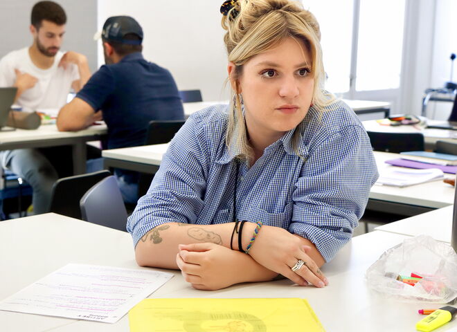 Eine Teilnehmerin sitzt an einem Tisch in einem Unterrichtsraum, hört aufmerksam zu und hat Unterlagen vor sich liegen.