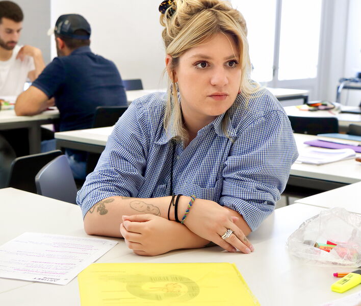 Eine Teilnehmerin sitzt an einem Tisch in einem Unterrichtsraum, hört aufmerksam zu und hat Unterlagen vor sich liegen.