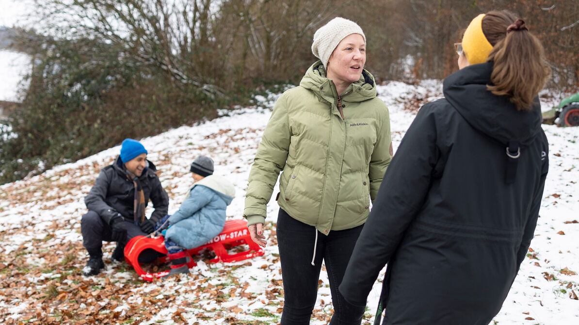 Zwei Erwachsene sprechen im Freien im Schnee; im Hintergrund spielen Kinder mit einem Schlitten.