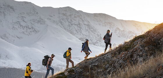 Cinq personnes montent un versant de montagne escarpé avec des sacs à dos, avec des montagnes enneigées en arrière-plan.