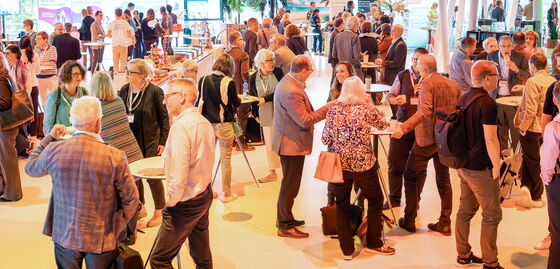 De nombreux participants au congrès se tiennent dans une grande salle autour de tables hautes et échangent entre eux.