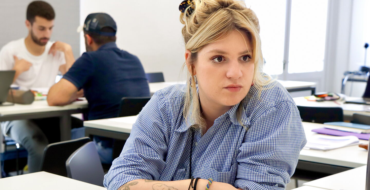 Eine Teilnehmerin sitzt an einem Tisch in einem Unterrichtsraum, hört aufmerksam zu und hat Unterlagen vor sich liegen.