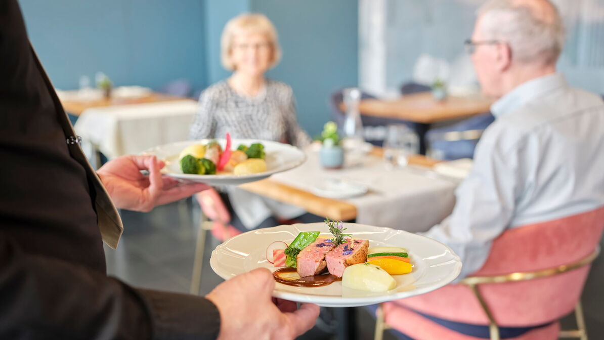 Une personne du service apporte deux assiettes de plats, tandis que deux personnes âgées sont assises à table et attendent leur repas.