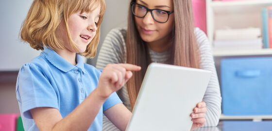 Une professionnelle de l’accompagnement est assise avec un enfant à une table, tandis que l’enfant montre quelque chose sur une tablette.