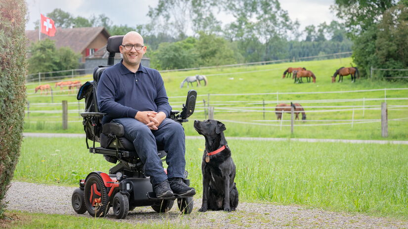 Matyas Sagi-Kiss sitzt im Rollstuhl in der Natur; neben ihm sitzt sein Assistenzhund.