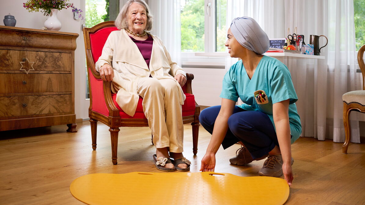 Une femme âgée est assise en souriant dans un fauteuil, tandis qu’une professionnelle des soins est accroupie à côté d’elle et ajuste un tapis jaune posé au sol.